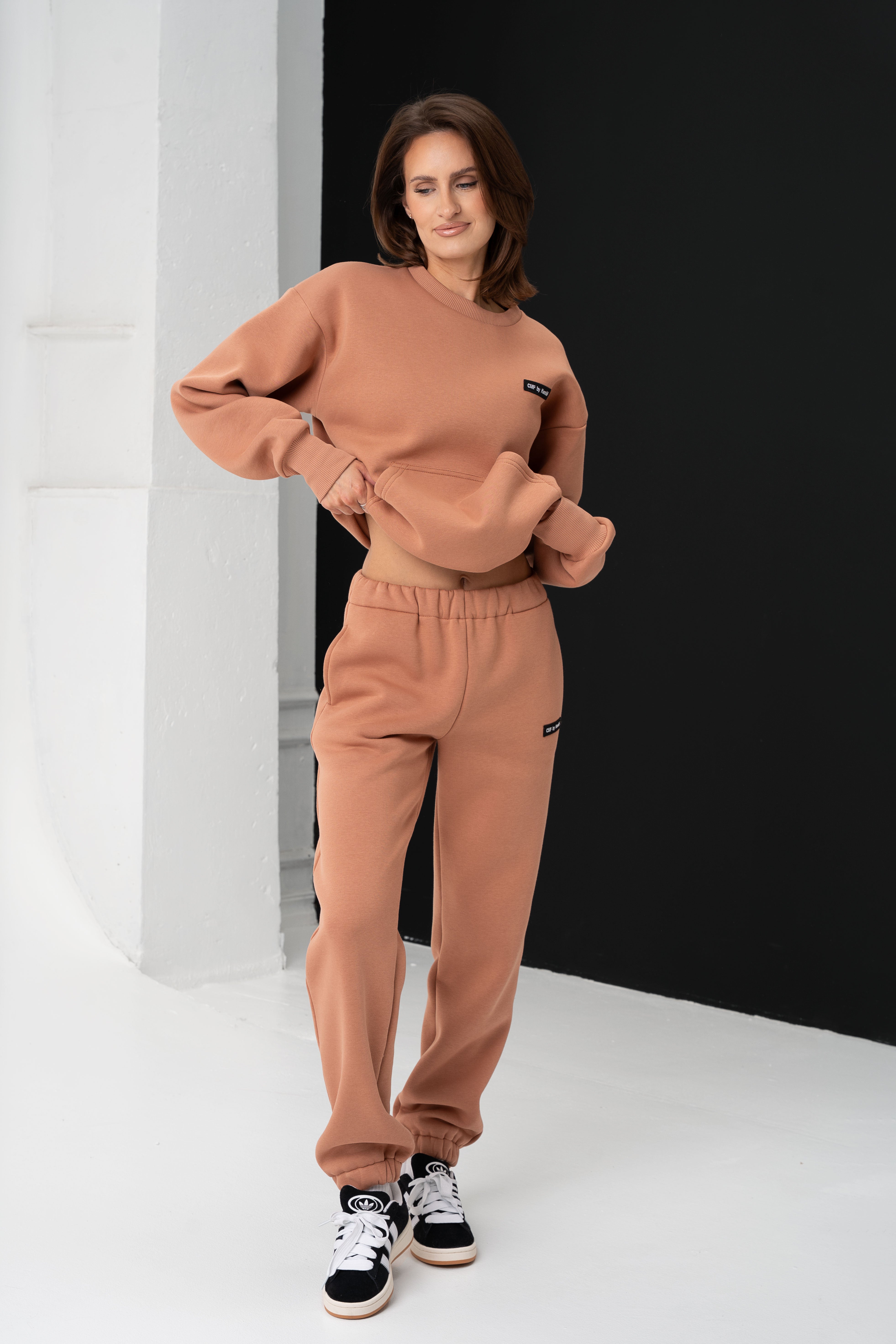 Moteris dėvėdama cmf by kamilė laisvalaikio treningus, lengvai pakelia mocha-mousse spalvos džemperį, atskleisdama jo laisvą siluetą.