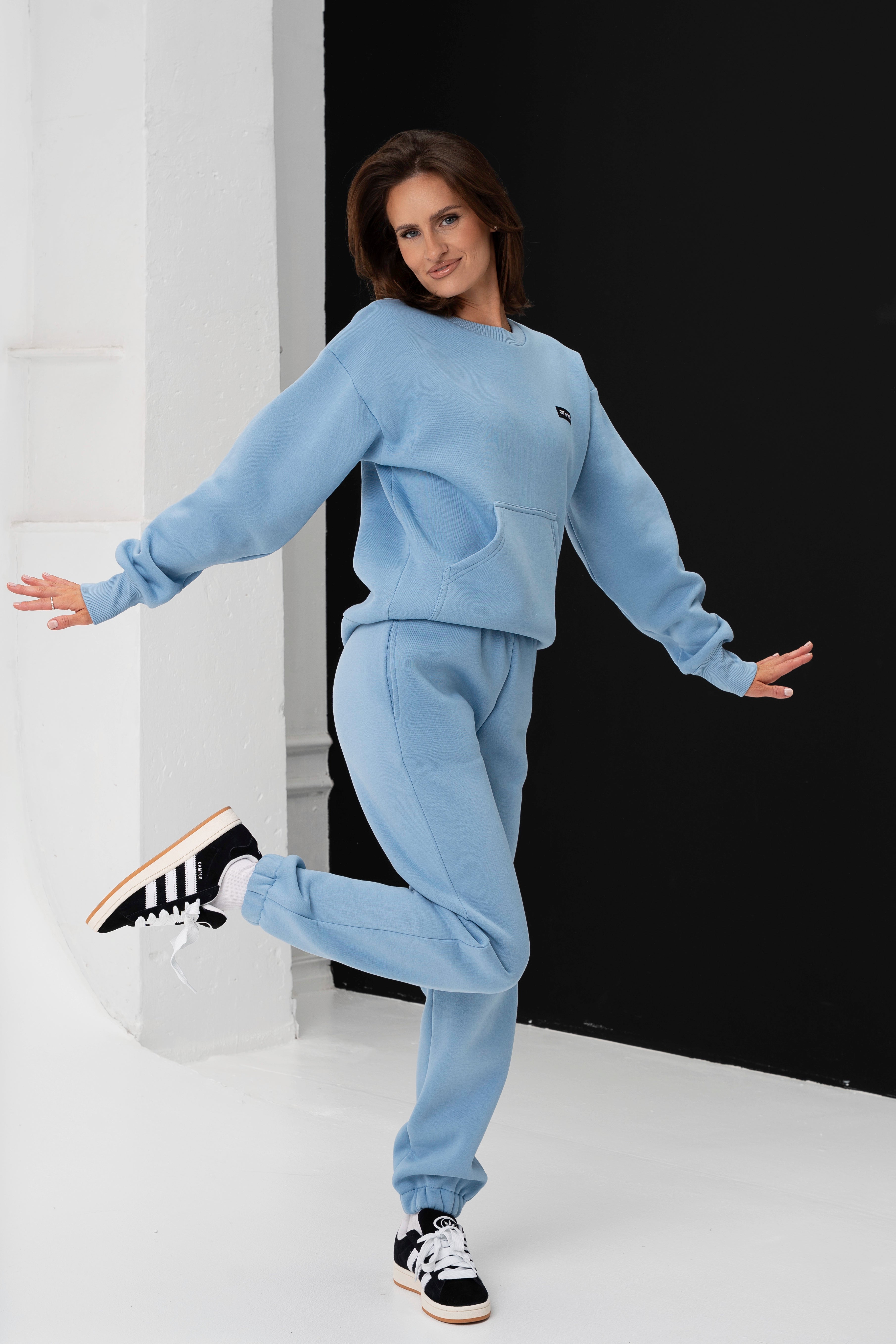 Moteris mėlynais treningais šoka pakėlusi vieną koją į orą, plačiai šypsosi