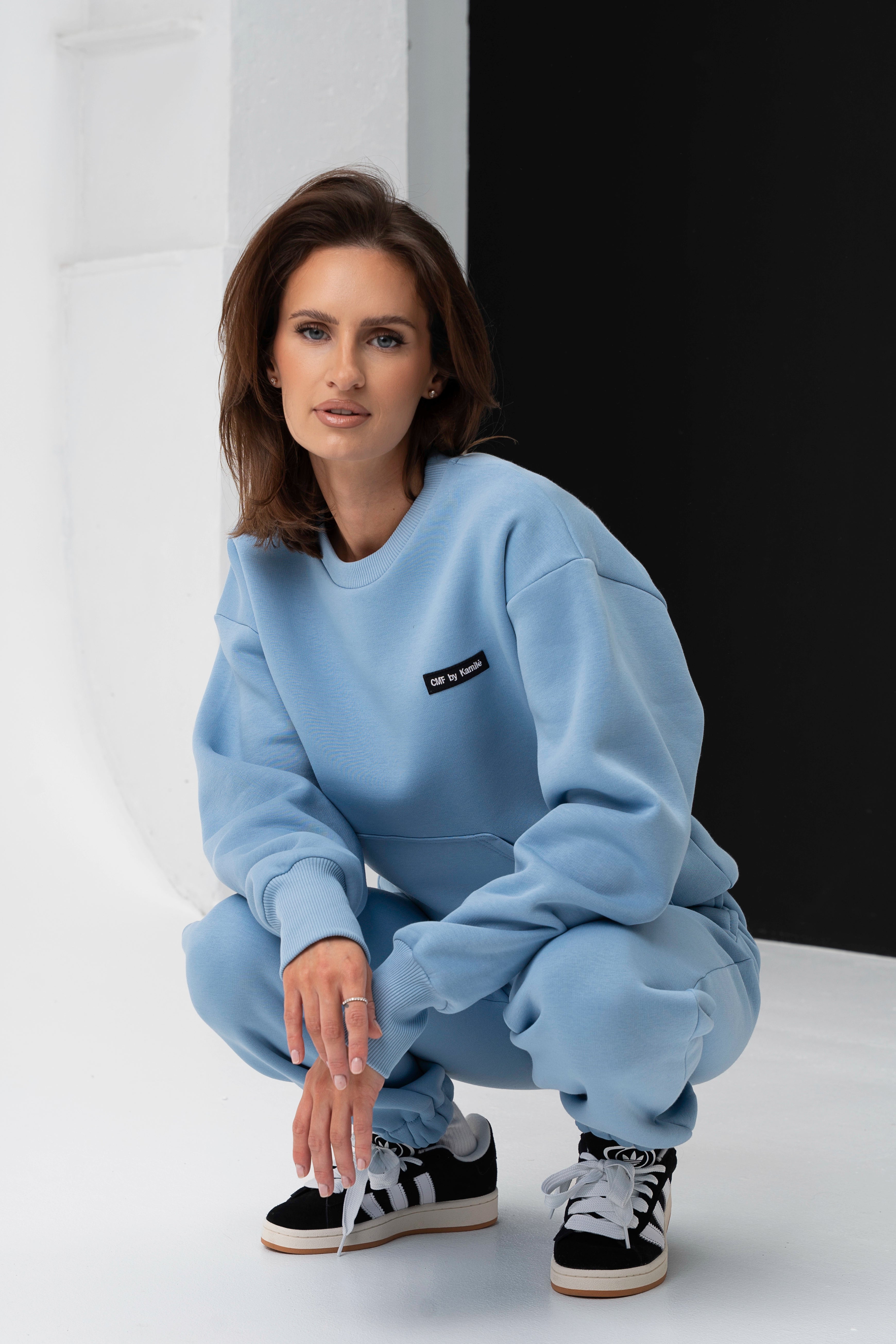Moteris pritūpusi dėvi mėlynus laisvalaikio treningus, dėmesys sutelktas į džemperio logotipą CMF by Kamilė