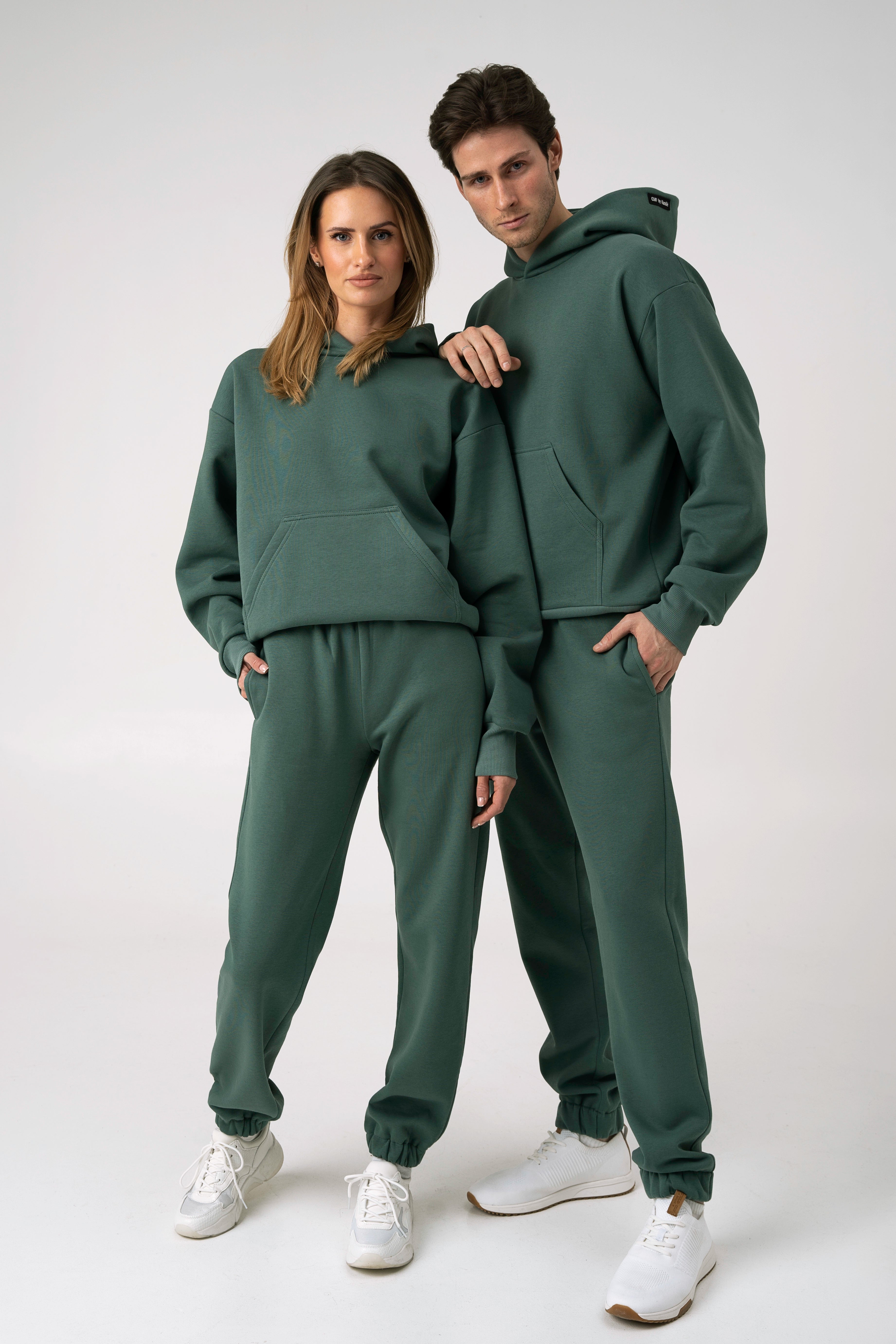Vyras ir moteris dėvi derančius žalius laisvalaikio treningus – džemperius su kapišonais ir kelnes su kišenėmis. Jie pozuoja kartu, stovėdami neutralios spalvos fone.