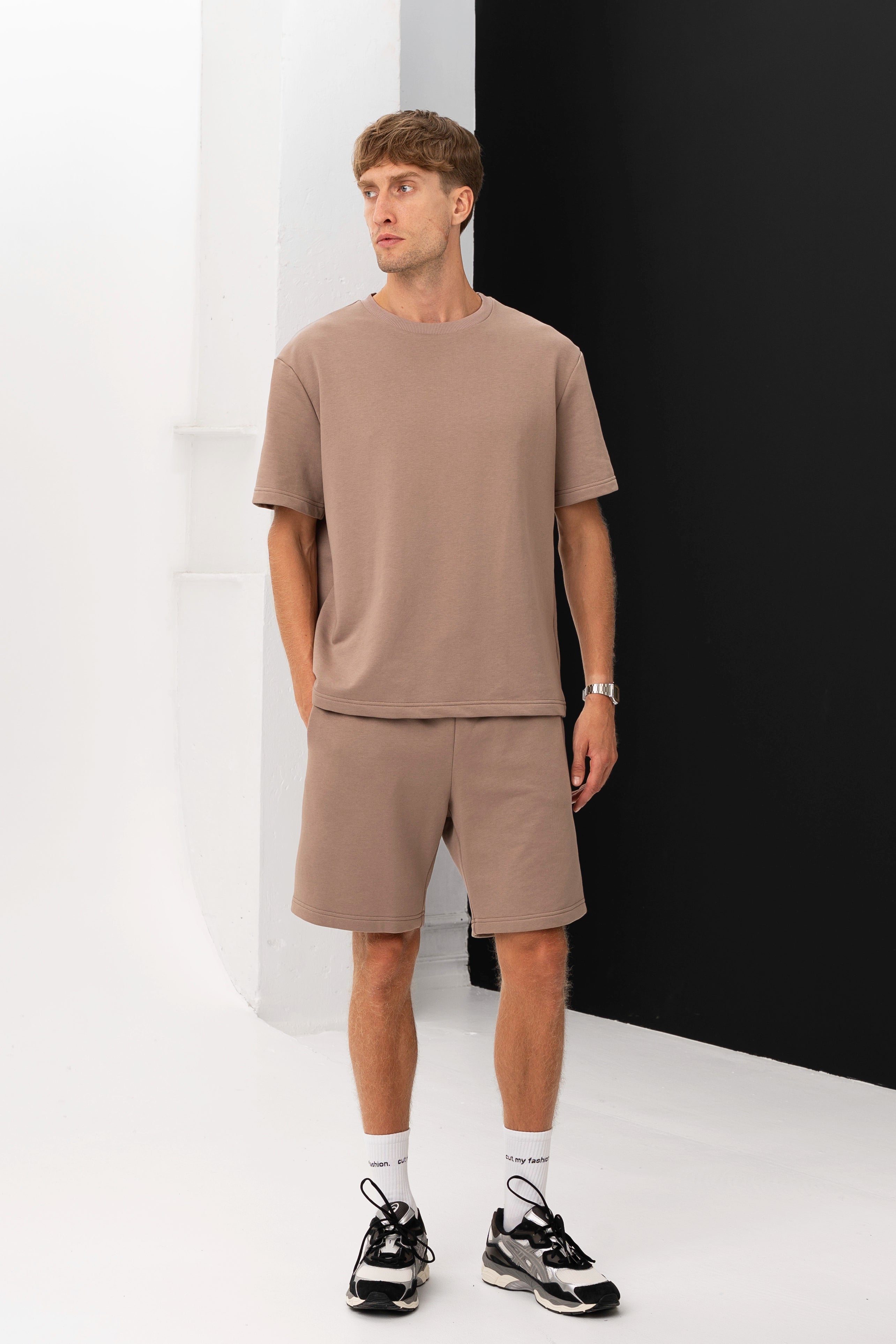 Modelis su rudais marškinėliais ir šortais pozuoja minimalistinėje studijoje, rankas laikydamas kišenėse