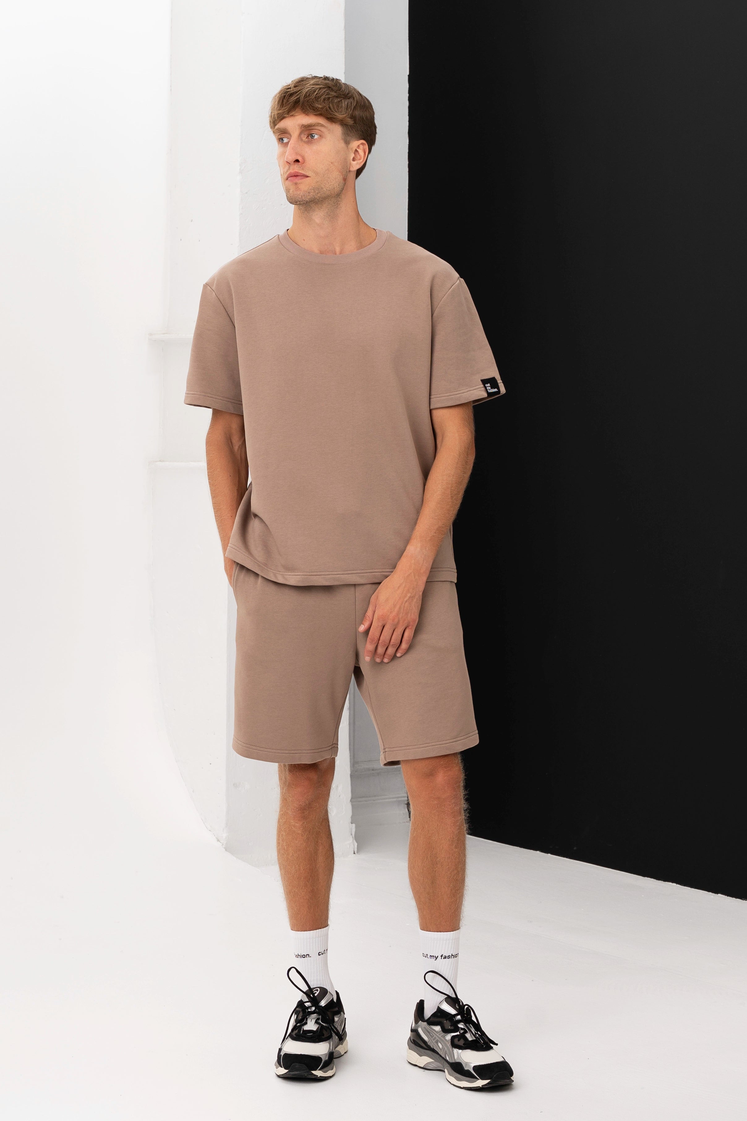 Vyras dėvi rudos spalvos marškinėlius ir šortus, stovėdamas prie baltos sienos su juoda detale