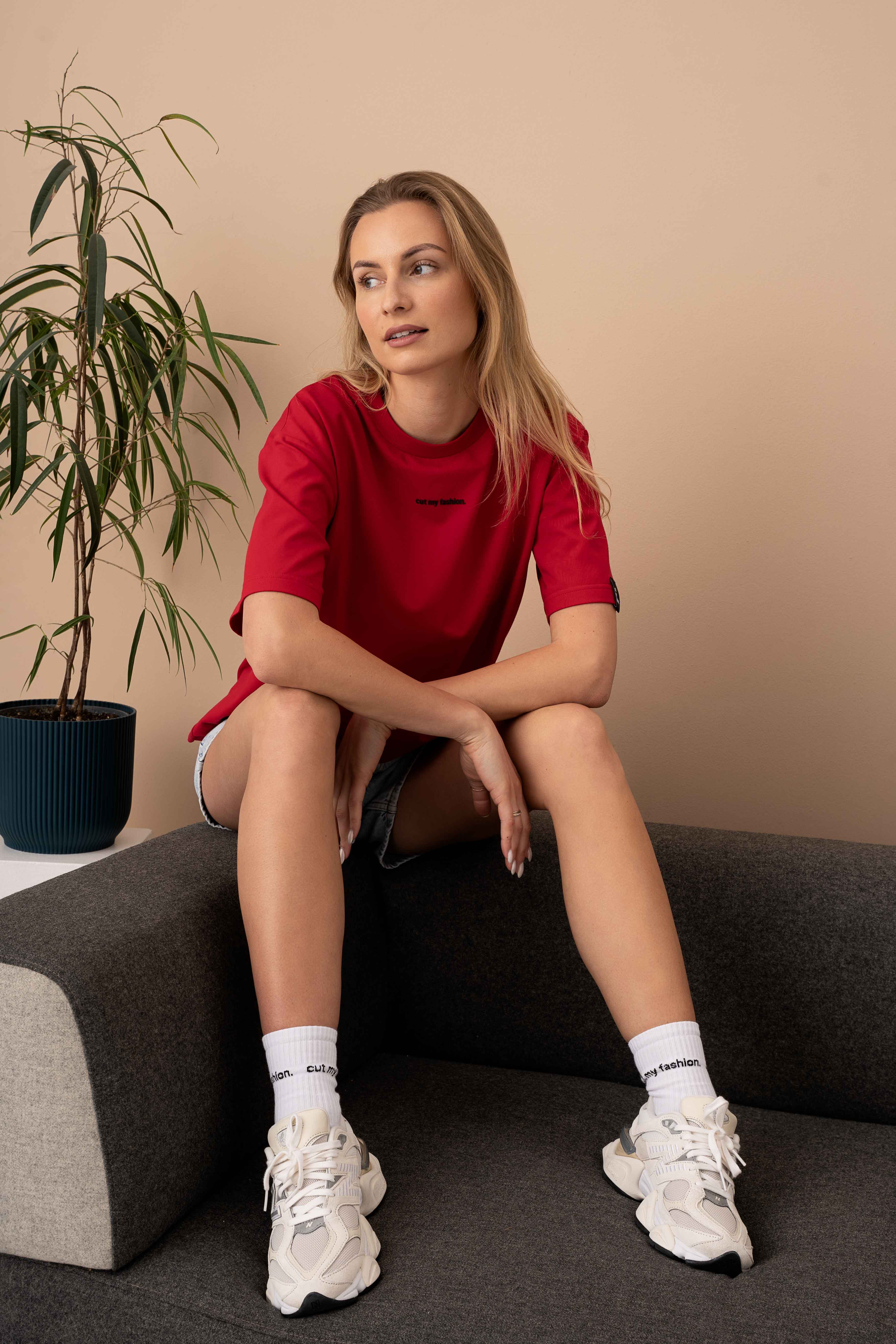 modelis su ryškiai raudonais oversized marškinėliais ir džinsiniais šortais, sėdi ant sofos, žiūri į šoną