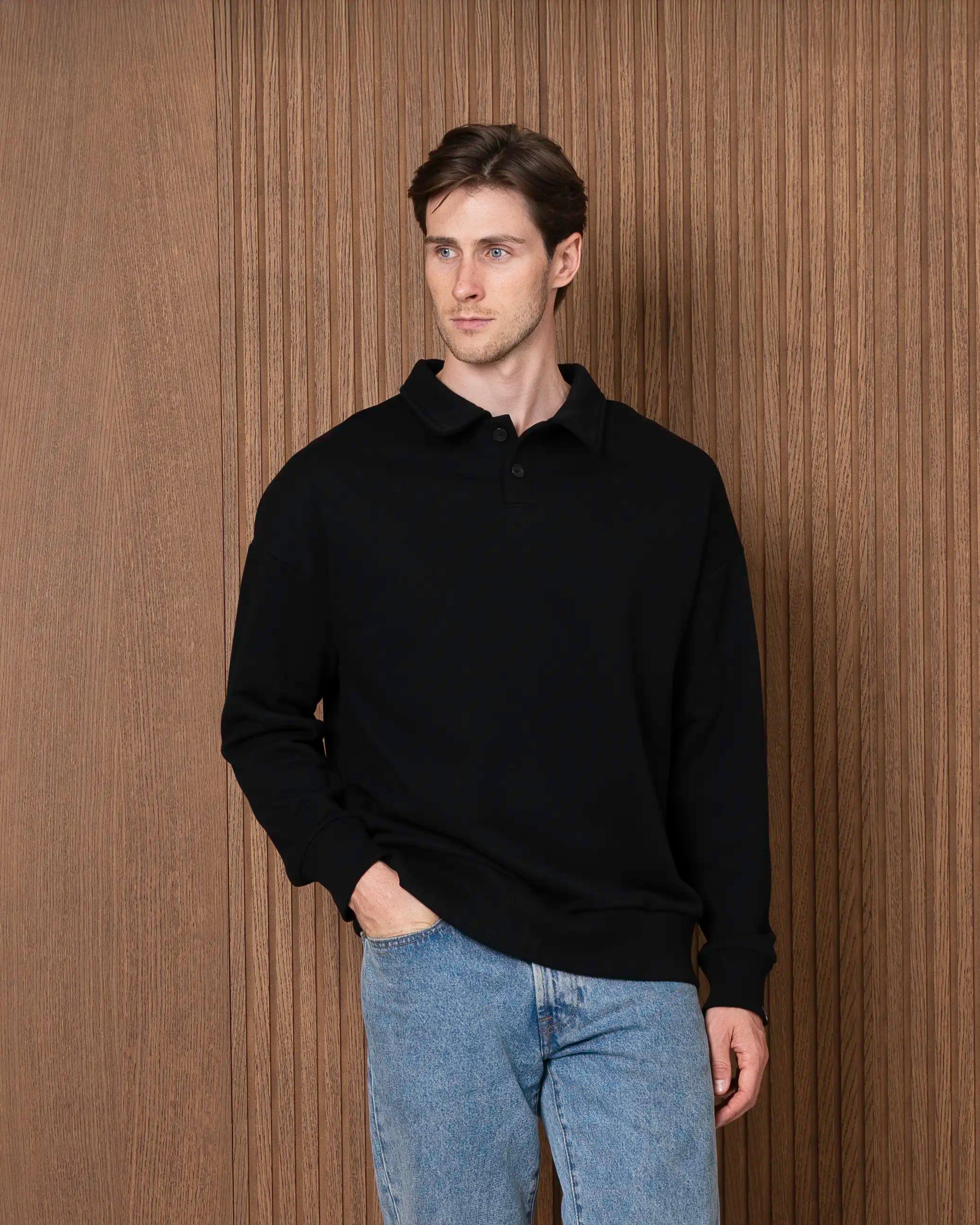 Vyras vilki juodą oversized džemperį su polo tipo apykakle užsegama sagomis, derintą su mėlynais džinsais.
