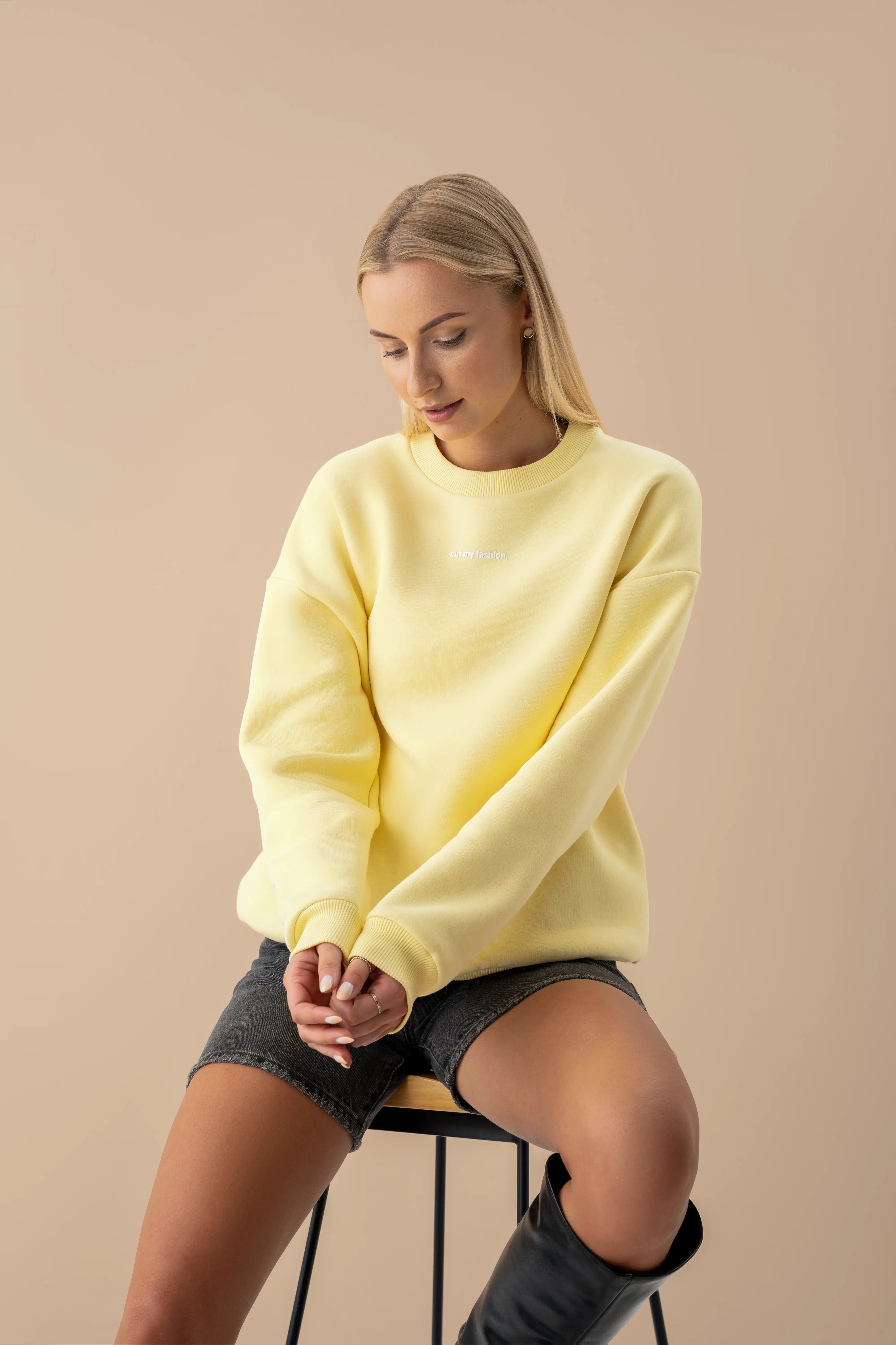geltonas oversized cutmyfashion džemperis iš OEKO TEX medvilnės su pūkeliu modelis sėdi ant kėdės vilki juodus šortus studijos aplinkoje pagaminta Lietuvoje