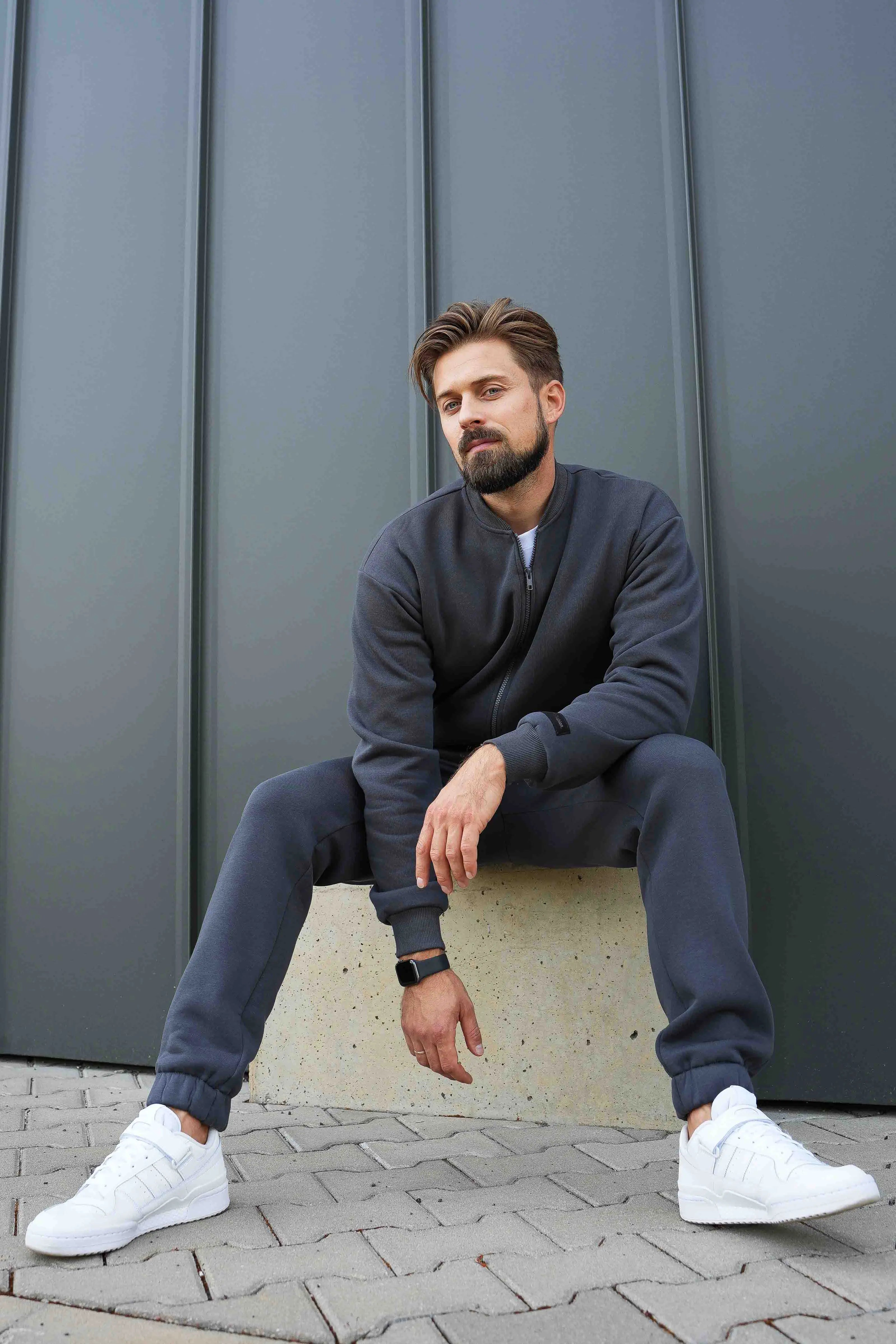vyras su cutmyfashion tamsiais treningais sėdi ant betoninės pakylos, žiūri į objektyvą