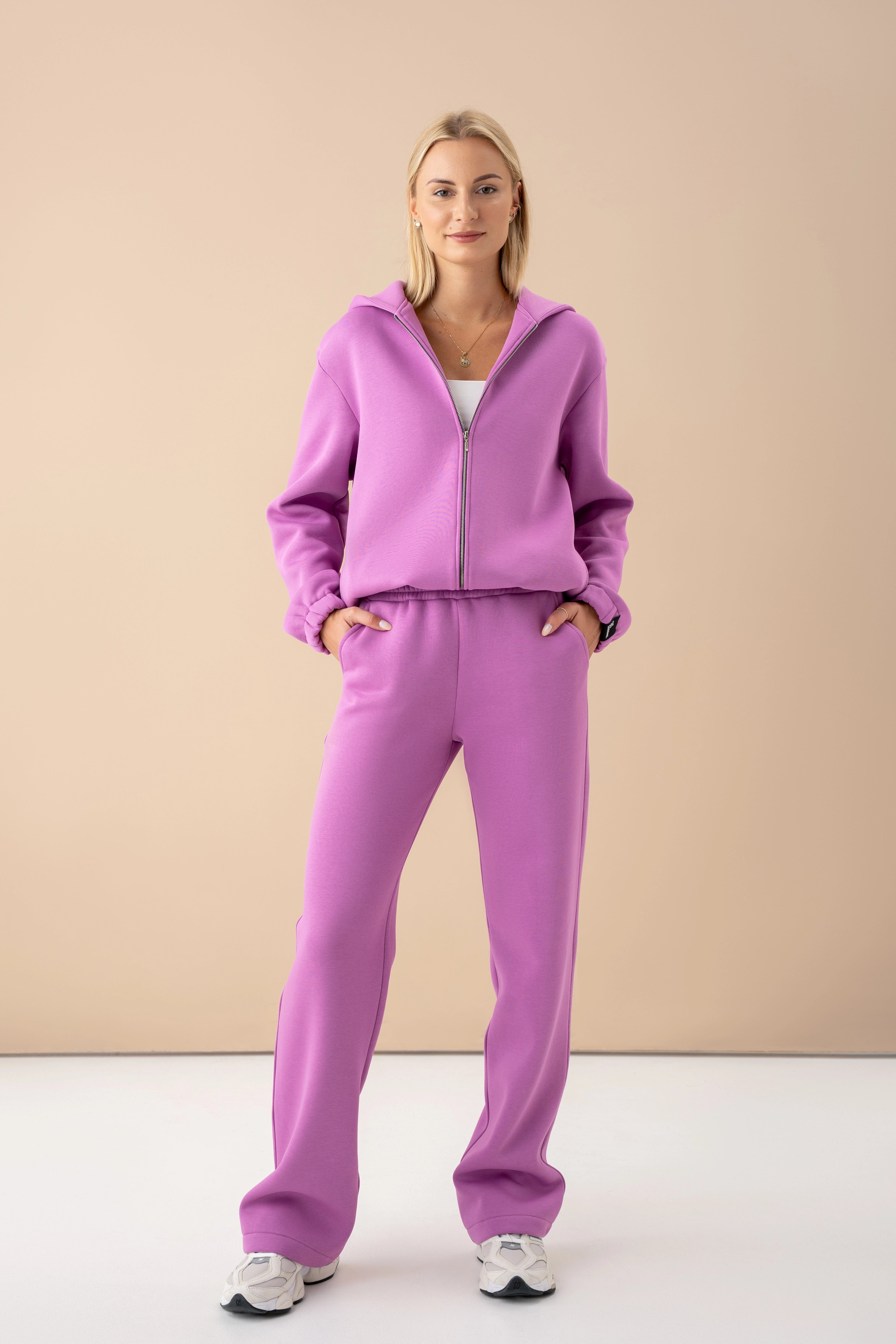 moteris vilki šaltibarščių violetinius cutmyfashion treningus, stovi studijos fone, rankas įkišusi į kišenes