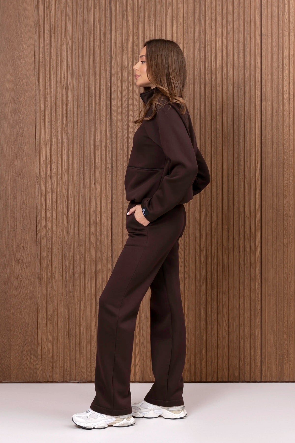 Šokolado spalvos laisvalaikio kostiumas iš šono, matomas džemperio siluetas ir tiesus kelnių kirpimas.