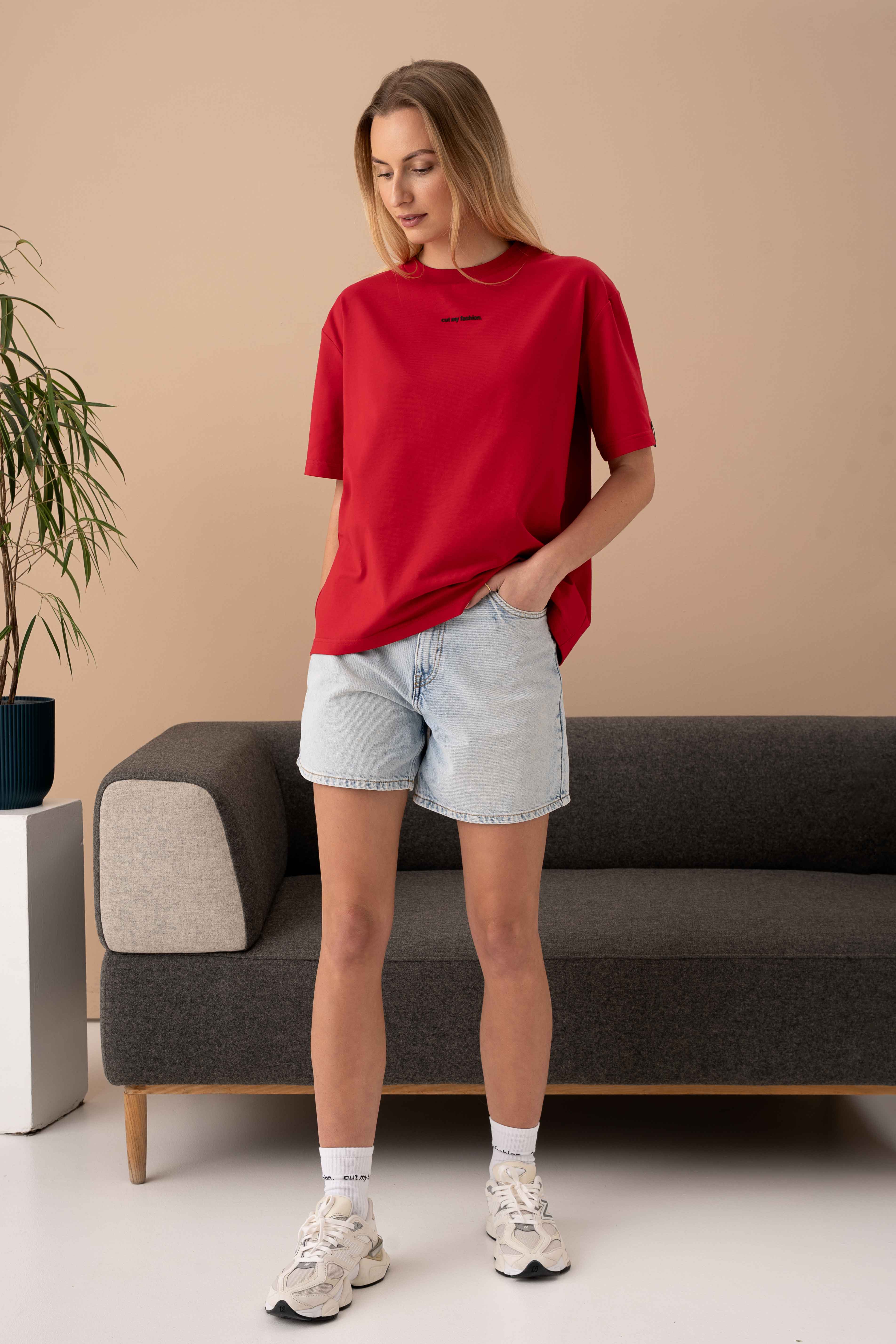moteris vilki raudonus cutmyfashion oversized marškinėlius ir džinsinius šortus, stovi prie sofos studijoje
