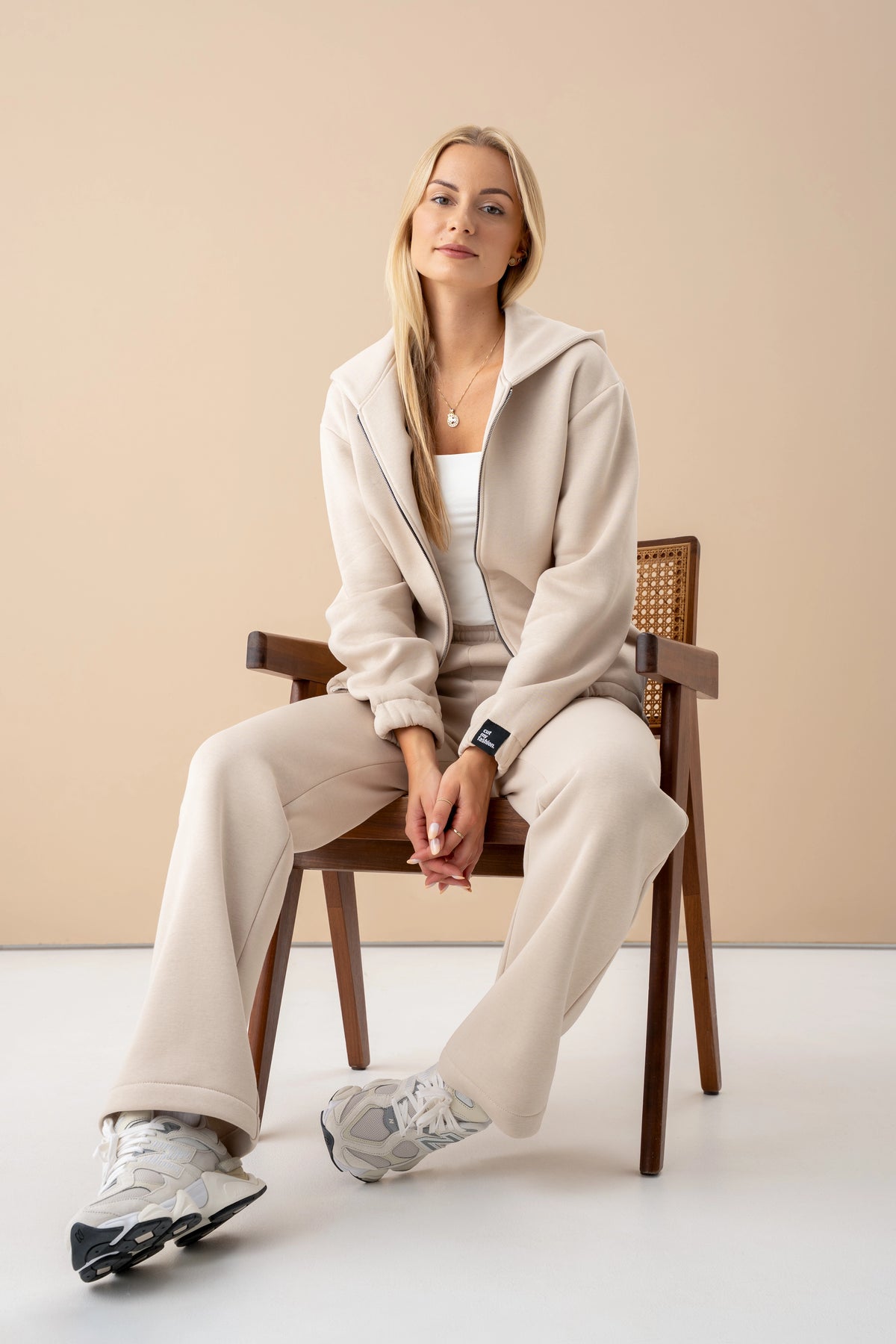Ant medinės kėdės sėdintis modelis vilki šviesiai smėlinį komplektą; matomi paraukti rankogaliai ir juoda cutmyfashion etiketė.
